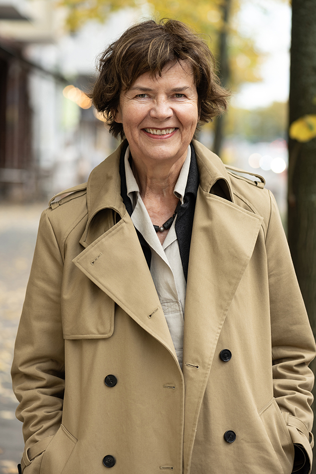 Anne Lampen im beigen Trenchcoat auf einer herbstlichen Straße