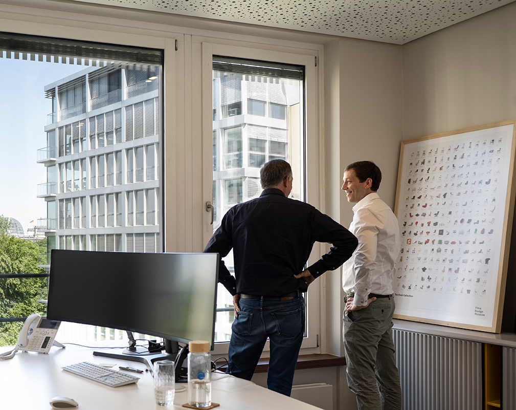 Zwei Männer im Gespräch am Fenster eines Büros mit Blick auf die Nachbargebäude