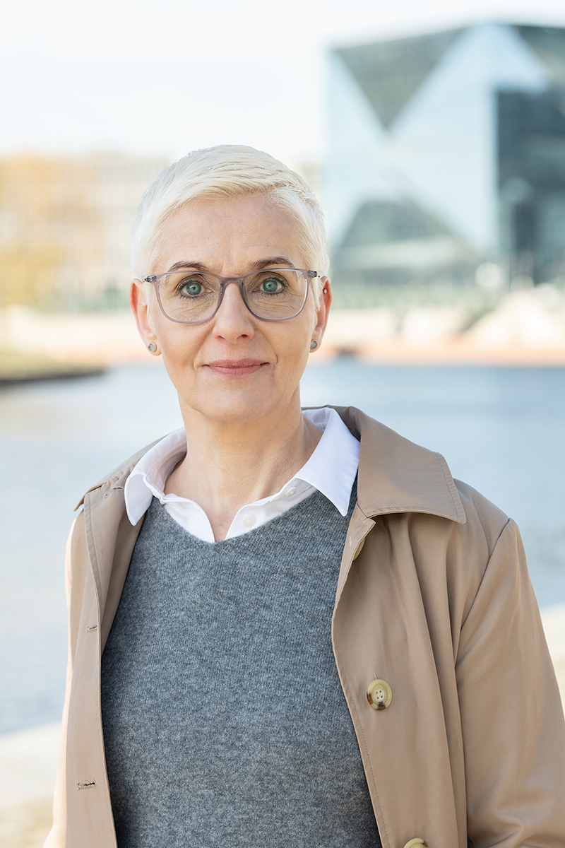 Porträt von Kati Fink mit Brille im Trenchcoat an der Spree