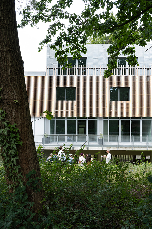 Wohnanlage mit Holzlamellen-Fassade, im Vordergrund Baumstamm und Gruppe von Menschen im Garten